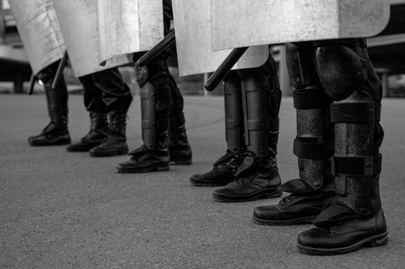 Image en noir et blanc de policiers anti-émeute debout en ligne, tenant des boucliers, portant des équipements de protection et des bottes sur une surface en béton.