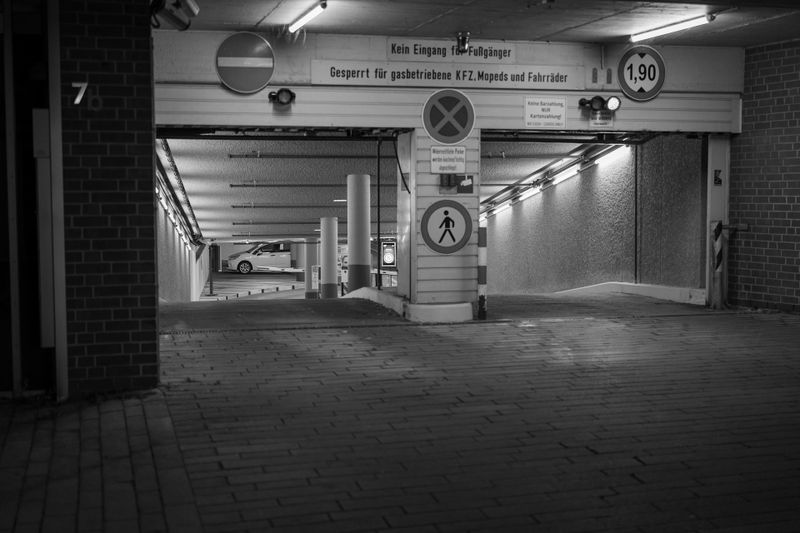 Image en noir et blanc de l’entrée d’un parking souterrain, avec une limitation de hauteur et des panneaux piétons, ainsi qu’une voiture stationnée visible à l’intérieur.