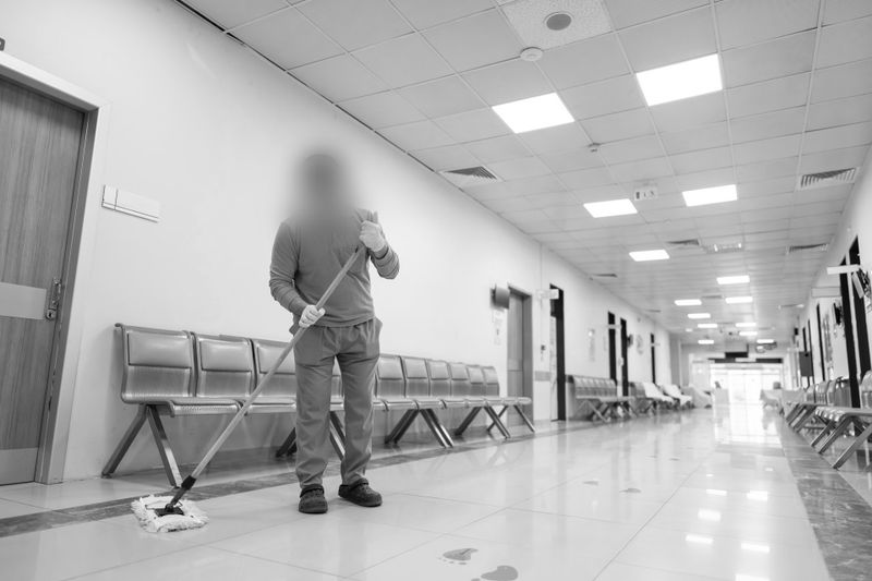 Una persona fregando el suelo en el pasillo de un hospital con sillas vacías alineadas en las paredes.