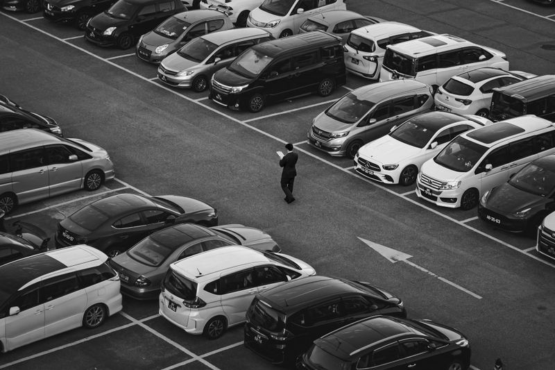 Vue aérienne d'un parking avec des rangées de voitures garées. Une personne marche entre les voitures, un téléphone à la main. Image en noir et blanc.