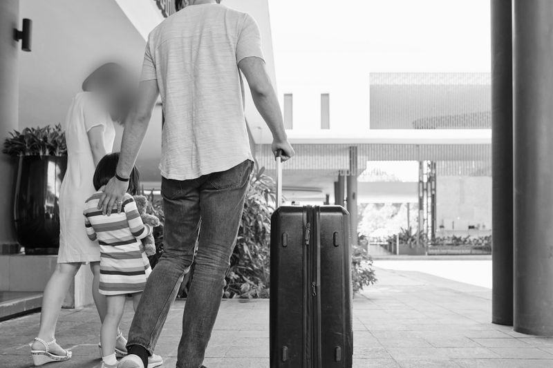 Une famille de trois personnes, avec un jeune enfant, avance avec une valise à roulettes dans un espace extérieur moderne. L’image est en noir et blanc.