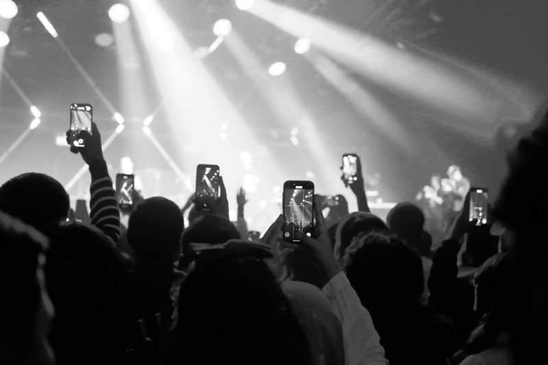 Crowd at a concert holding up smartphones, capturing the performance. Bright stage lights illuminate the scene in black and white.