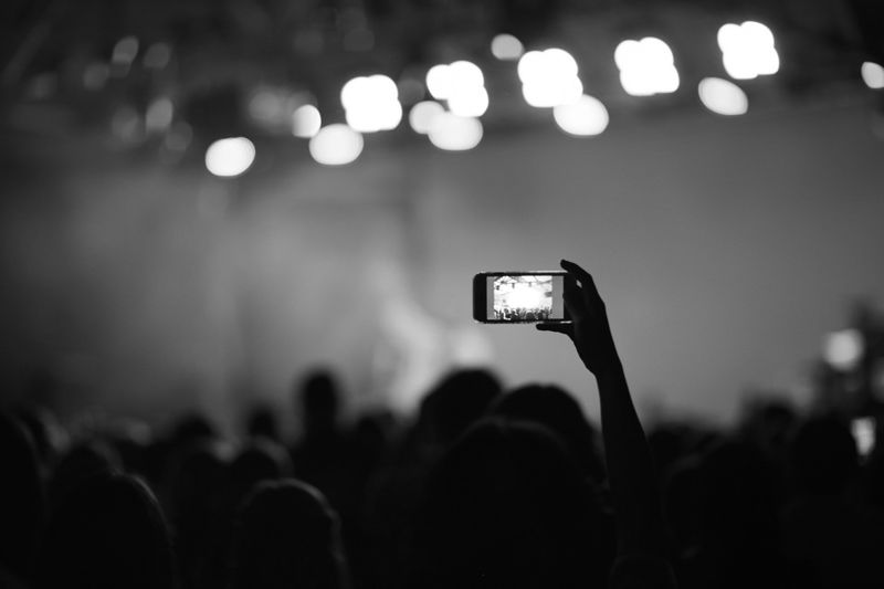 Black and white image of a crowd at a concert, with a hand holding a smartphone capturing the stage, blurred lights in the background.