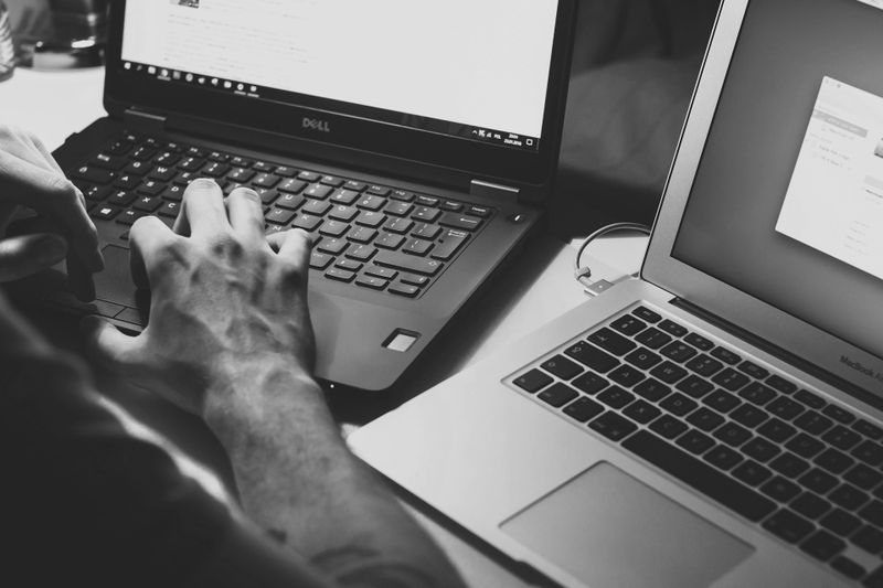 Une personne tape sur un ordinateur portable Dell à côté d'un MacBook ouvert, tous deux sur un bureau, dans une pièce faiblement éclairée.