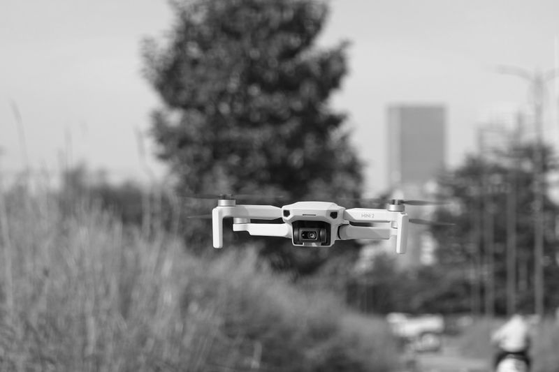 A small drone hovers outdoors in a grassy area, with blurred trees and buildings in the background. Black and white image.