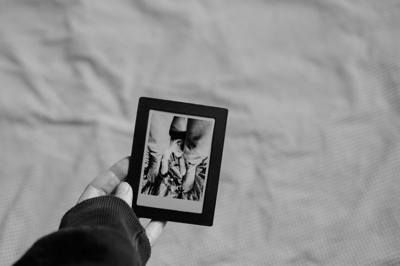 Une main tient une photo encadrée de noir, montrant un gros plan d'une personne en noir et blanc. L'arrière-plan est une surface de tissu floue.