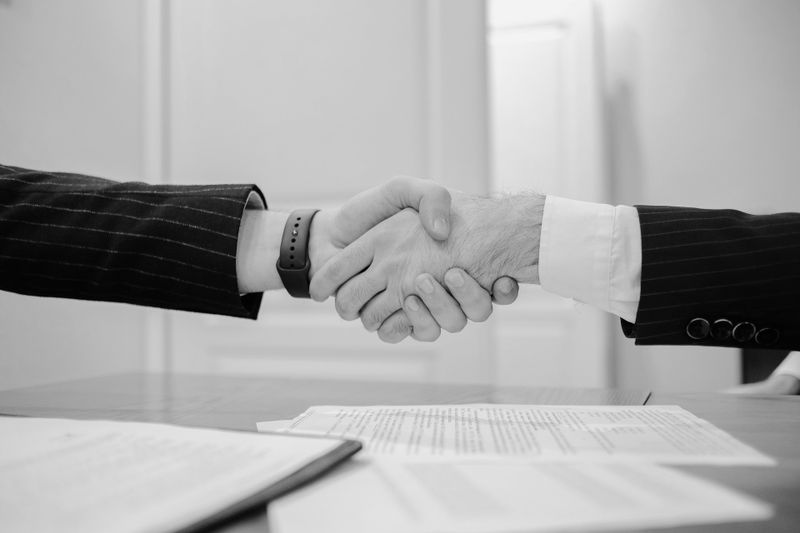 Two people in suits shaking hands over a table with documents, symbolizing a business deal or partnership.