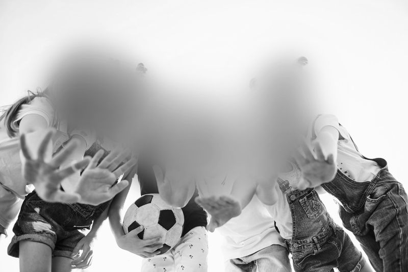 Group of children standing in a circle, viewed from below, holding a soccer ball. Their faces are intentionally blurred. Black and white image.