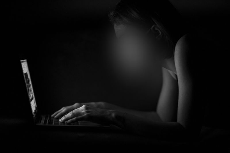 Personne dans une pièce sombre, visage flou, utilisant un ordinateur portable avec une lumière focalisée sur ses mains et son clavier.