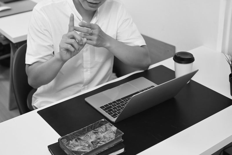 Une personne en chemise blanche fait des gestes, assise à un bureau avec un ordinateur portable, une tasse de café et des livres. Image en noir et blanc.