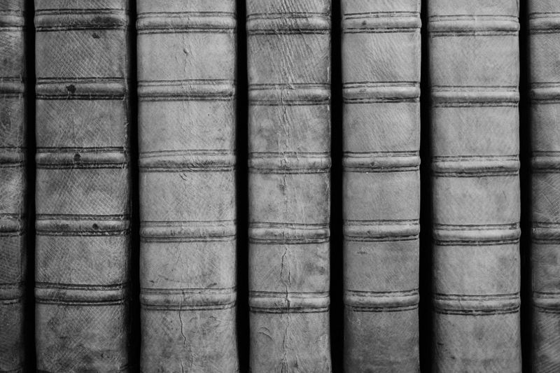 Gros plan de livres vintage reliés en cuir, alignés verticalement, montrant des dos texturés avec des crêtes horizontales, en noir et blanc.