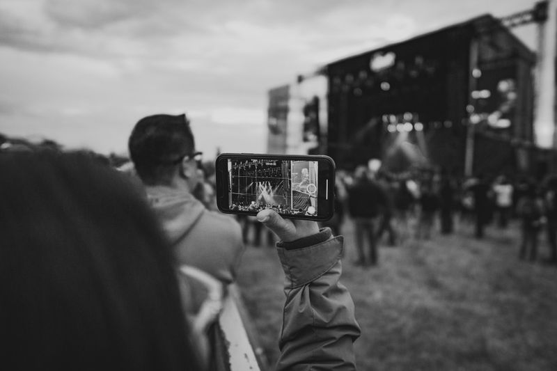 Person filming a concert stage with a smartphone in a crowd at an outdoor event, captured in black and white.