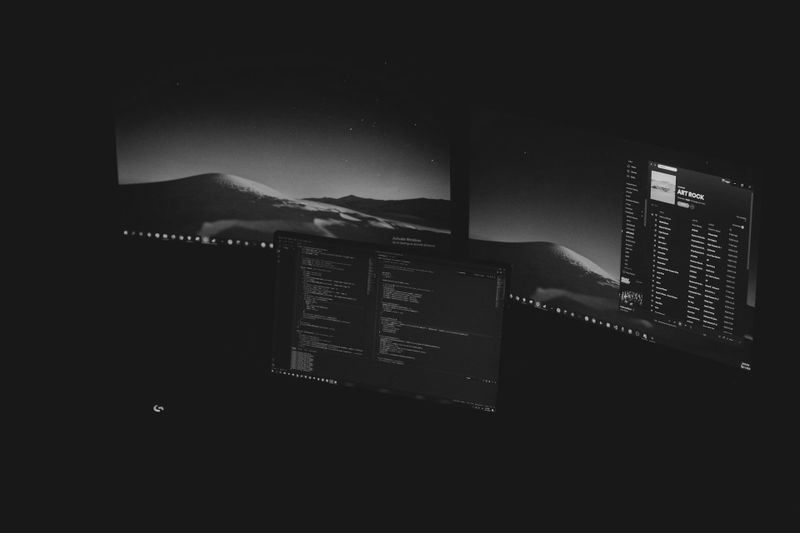 Three monitors display code and music playlists in a dark room. The screens show a desert landscape, code editor, and music app interface.