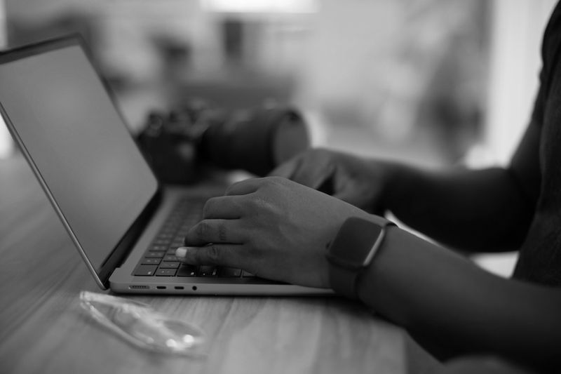 Une personne tape sur un ordinateur portable posé sur une table en bois, avec un appareil photo à proximité. Elle porte une montre connectée. Image en noir et blanc.