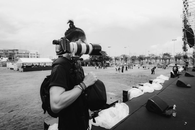 Photographer with a large camera lens stands on an outdoor stage, capturing a distant scene. Overcast sky and scattered people in the background.
