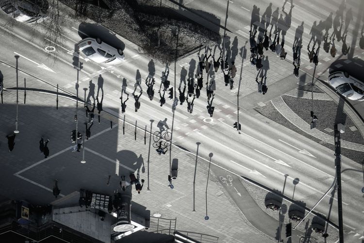 Widok z lotu ptaka na ludzi spacerujących i jeżdżących na rowerach po miejskiej ulicy, rzucających długie cienie w słoneczny dzień.