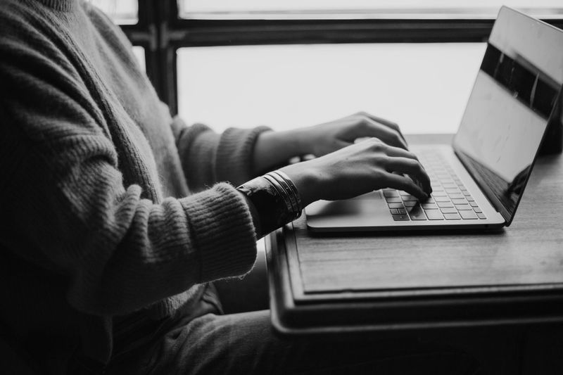 Person typing on a laptop at a wooden table, wearing a sweater. Black and white image with natural light from a window.