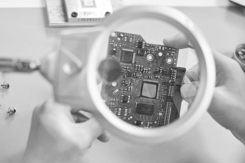 Mains tenant un circuit imprimé sous une loupe, se concentrant sur les composants électroniques dans une image en niveaux de gris.
