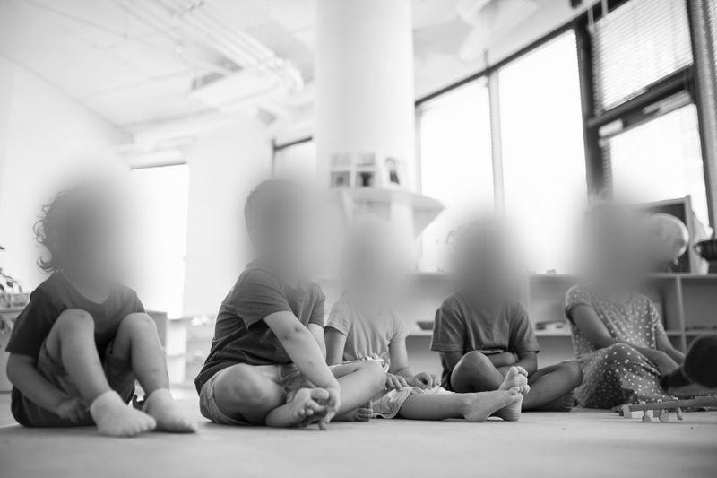 Des enfants assis en tailleur dans une salle de classe lumineuse, les visages floutés, suggérant une mise en valeur de l’environnement plutôt que des individus. Noir et blanc.