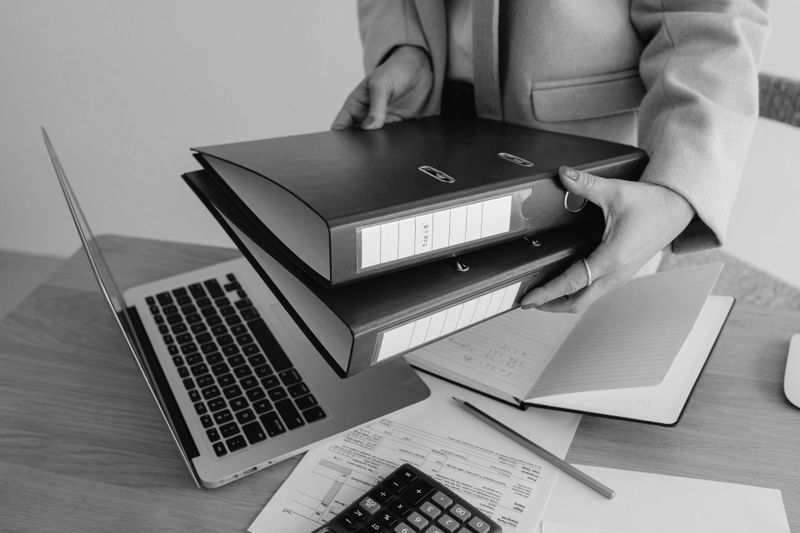 Person holding two large binders over a desk with a laptop, papers, and a calculator.