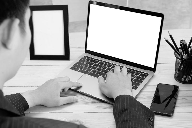 Personne en costume utilisant un ordinateur portable avec un écran vide à un bureau, à côté d'un smartphone, d'un pot à crayons et d'un cadre photo vide.