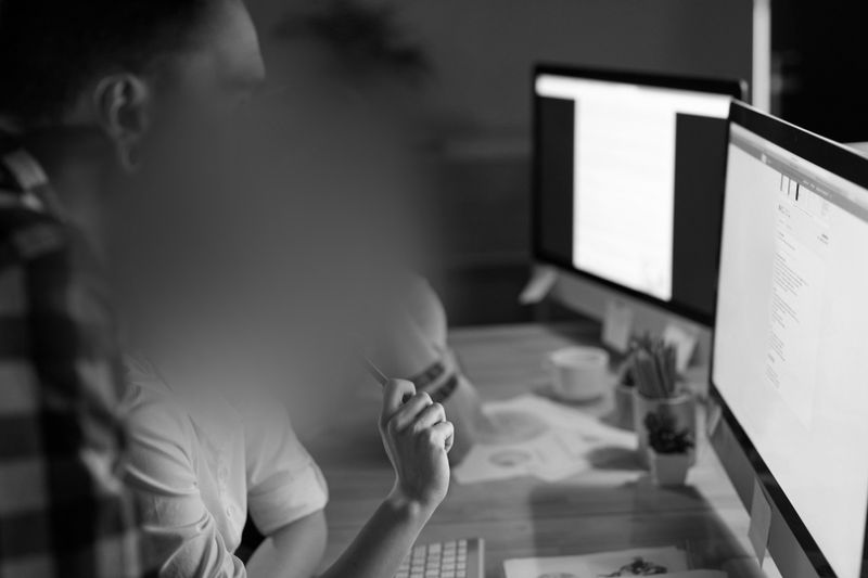 Two people are working at a desk with two monitors, focusing their gaze on a screen displaying text. The photo is black and white, and the people's faces are blurred.