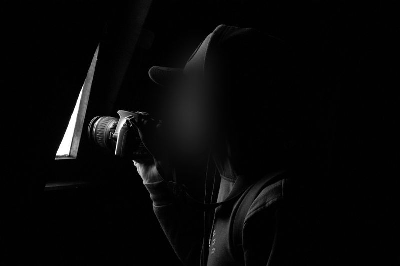 Une personne portant un sweat à capuche et une casquette tient un appareil photo et regarde à travers une fenêtre étroite dans un environnement sombre.