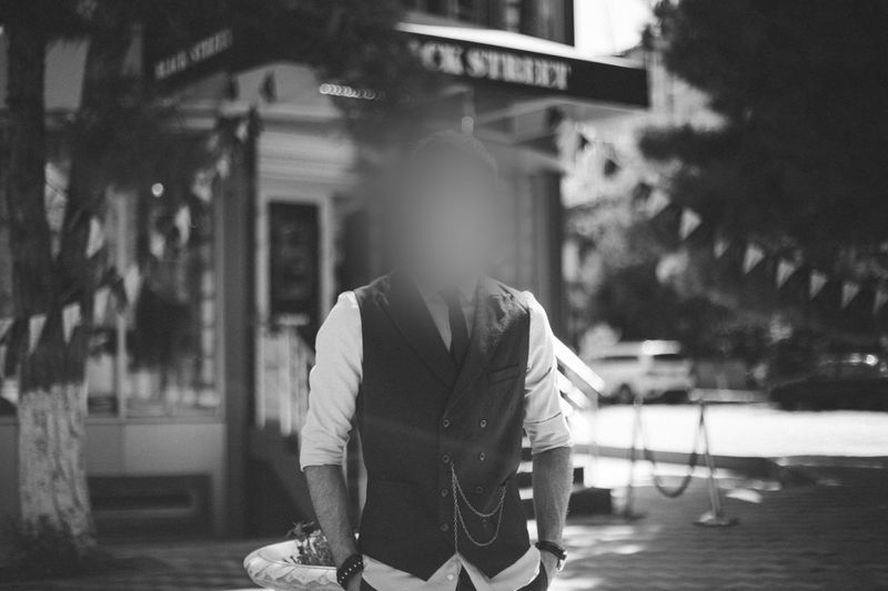 Une personne portant un gilet et une chemise blanche se tient dans une rue, avec son visage flou. Des arbres et un bâtiment sont en arrière-plan. Photo en noir et blanc.