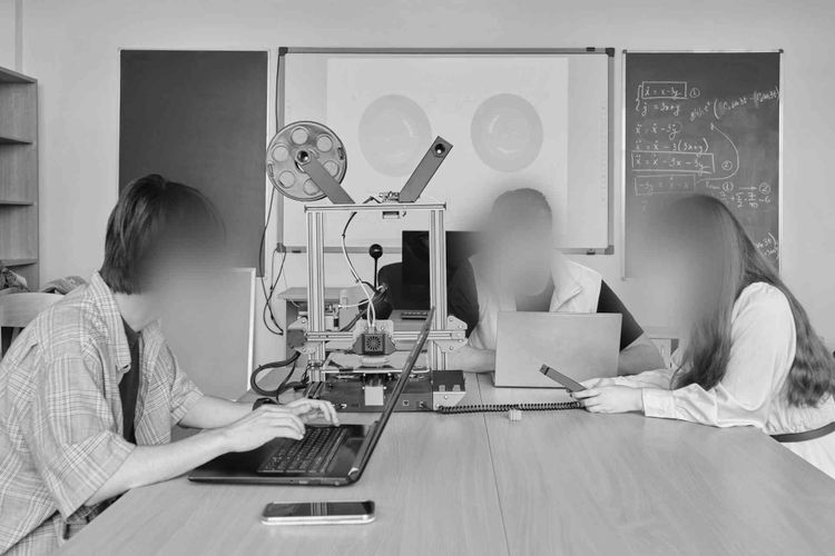 Trzy osoby pracujące na laptopach wokół drukarki 3D w klasie, w tle tablica i ekran projektora.