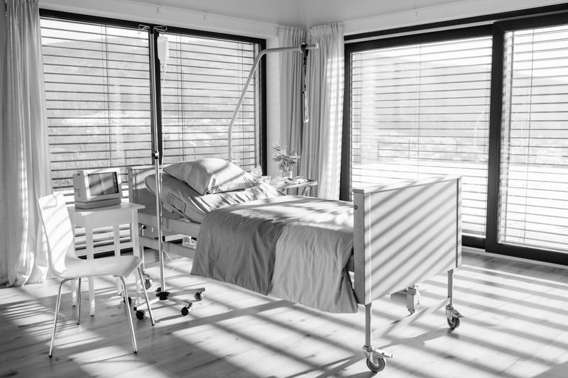 Habitación de hospital moderna con una cama, una silla y una tableta sobre un escritorio. Grandes ventanas con persianas dejan entrar mucha luz, proyectando sombras en el suelo.