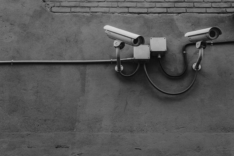 Deux caméras de surveillance montées sur un mur en béton texturé, reliées par des câbles apparents, surmonté d'une section en briques. Image en noir et blanc.