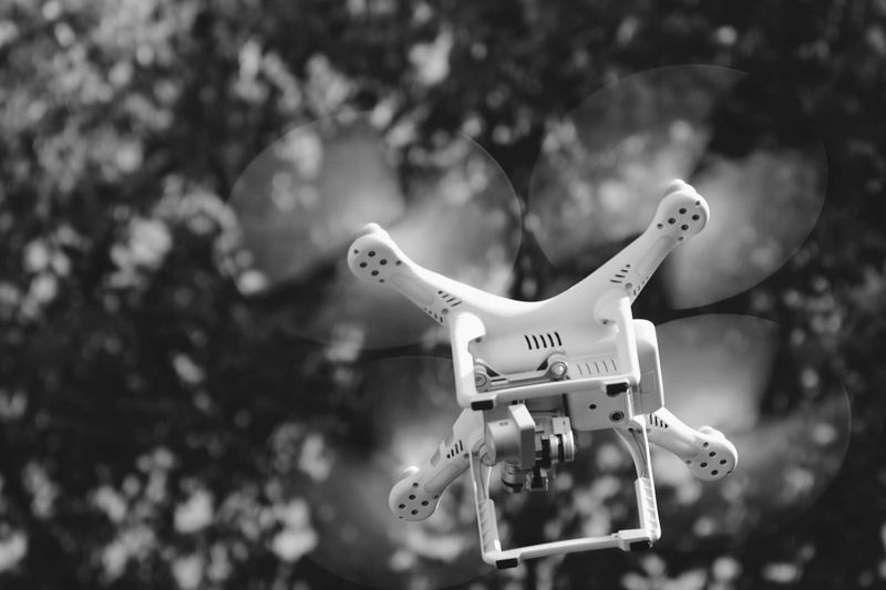 Black and white image of a quadcopter drone flying against a blurred background of trees.