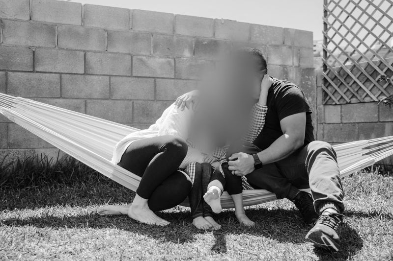 Famille assise sur un hamac dans un jardin, visages floutés, à côté d’un mur en brique par une journée ensoleillée. Photo en noir et blanc.