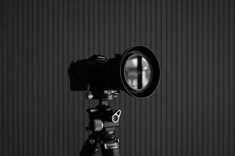 Una cámara profesional con un gran objetivo montado en un trípode frente a un fondo oscuro con rayas verticales.