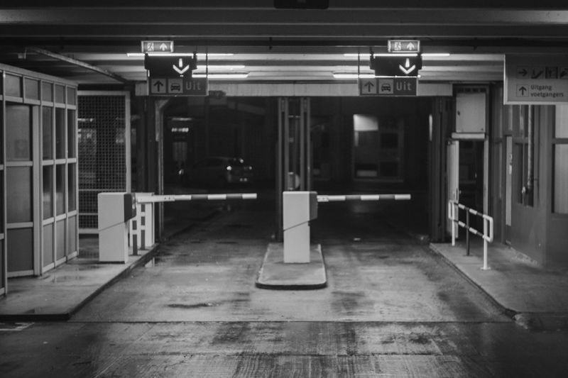 Image en noir et blanc de la sortie d’un parking vide, avec des barrières fermées et des panneaux de sortie éclairés au-dessus.