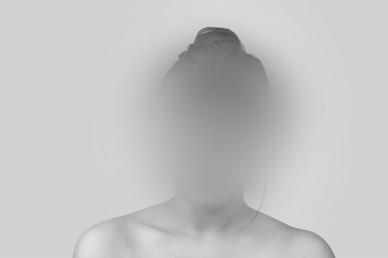 Blurred image of a person with an indistinct face against a gray background, hair tied up, and shoulders visible.