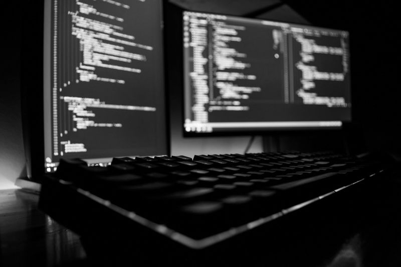 Primer plano en blanco y negro de un teclado con dos monitores que muestran código de programación en un espacio de trabajo oscuro.