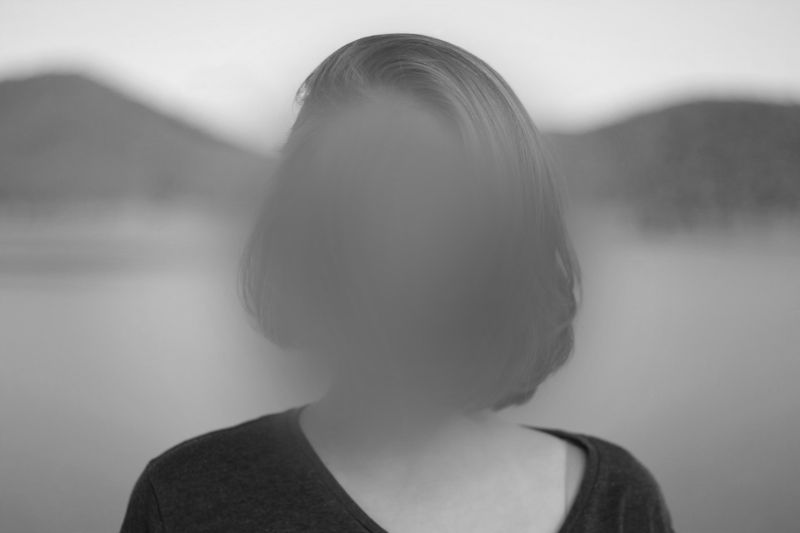 Black-and-white photo of a person with chin-length hair and a blurred face standing by a calm lake with hills in the background.