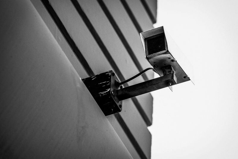 Black and white image of a security camera mounted on a building corner, angled downward, with a clear sky in the background.