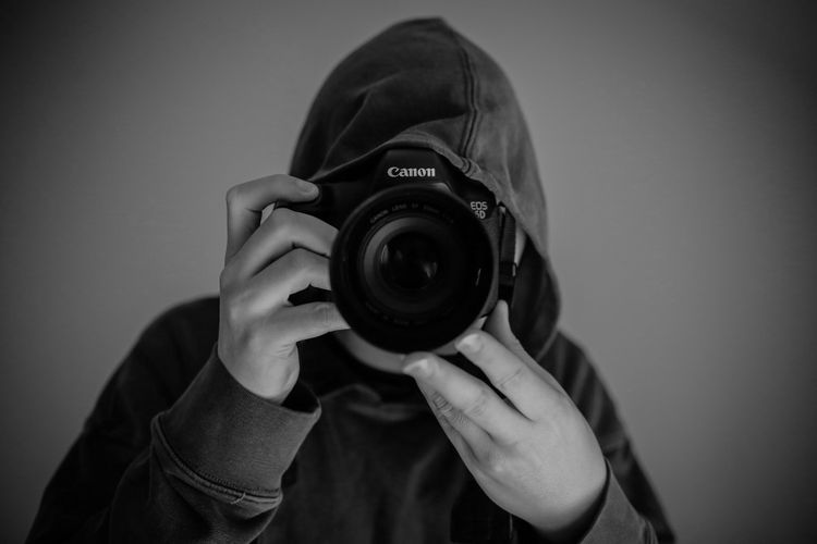 Osoba w bluzie z kapturem, trzymająca aparat Canon, robiąca zdjęcie. Zdjęcie jest czarno-białe.