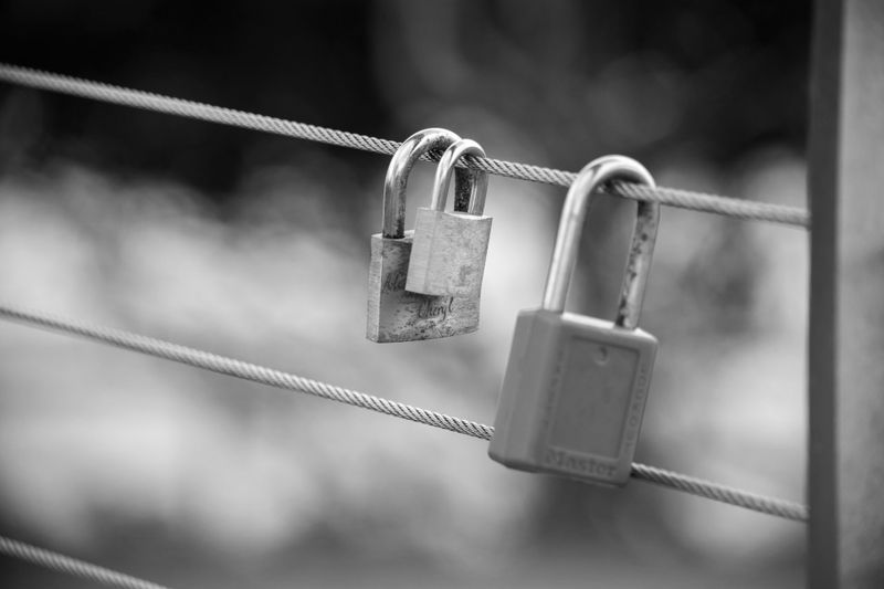 Two padlocks attached to parallel metal cables, with a blurred background in black and white.