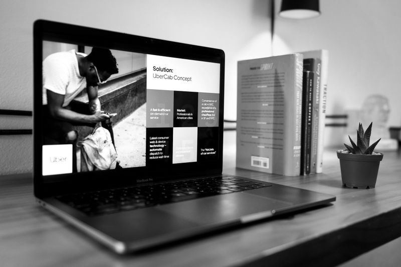 A black-and-white photo of a laptop on a desk displaying a presentation titled "UberCab Concept" on its screen, with books and a small plant nearby.