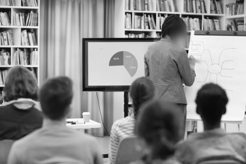 foto en blanco y negro que muestra una conferencia para jóvenes en una biblioteca, el ponente dibuja un mapa mental en la pizarra, tiene la cara borrosa