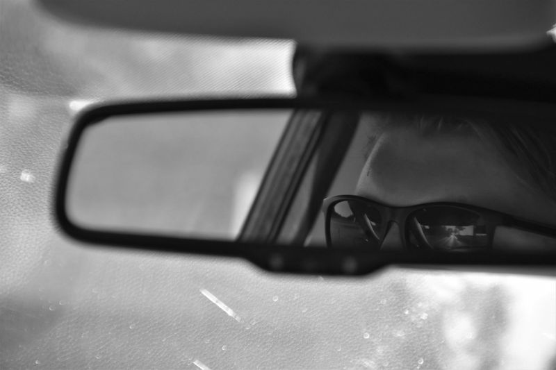 foto en blanco y negro desde el interior del coche, vista del espejo retrovisor en el que se refleja el rostro de un hombre con gafas de sol