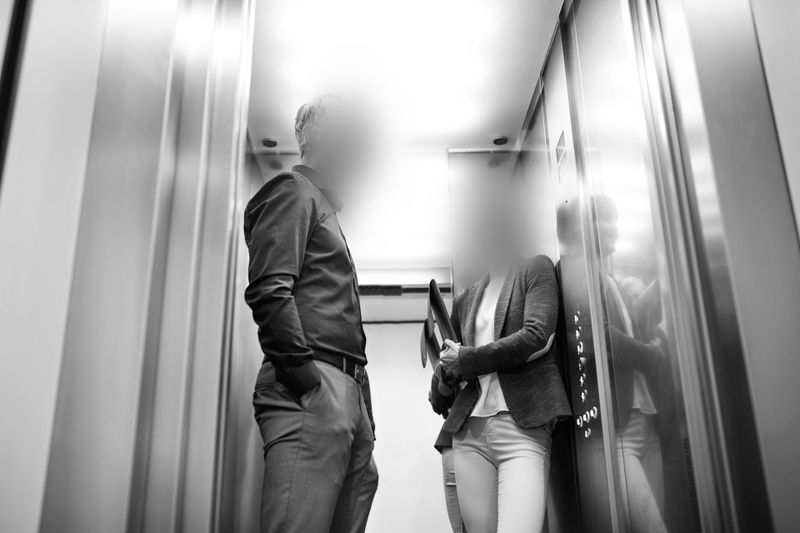 Two blurred individuals conversing in an elevator, one holding a folder. The image is black and white, capturing a reflective interior.