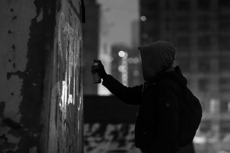 Silhouette of a person in a hooded jacket spray painting graffiti on a wall at night, with an urban cityscape in the background.