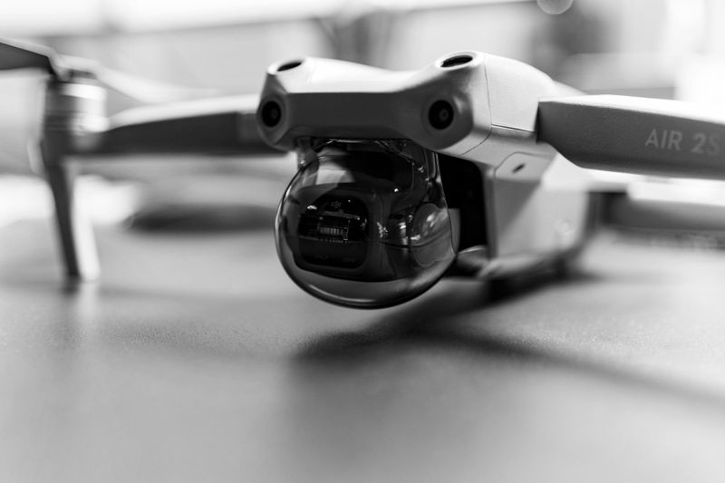 Close-up of a drone's camera in black and white, showing the lens and part of the body, resting on a flat surface.