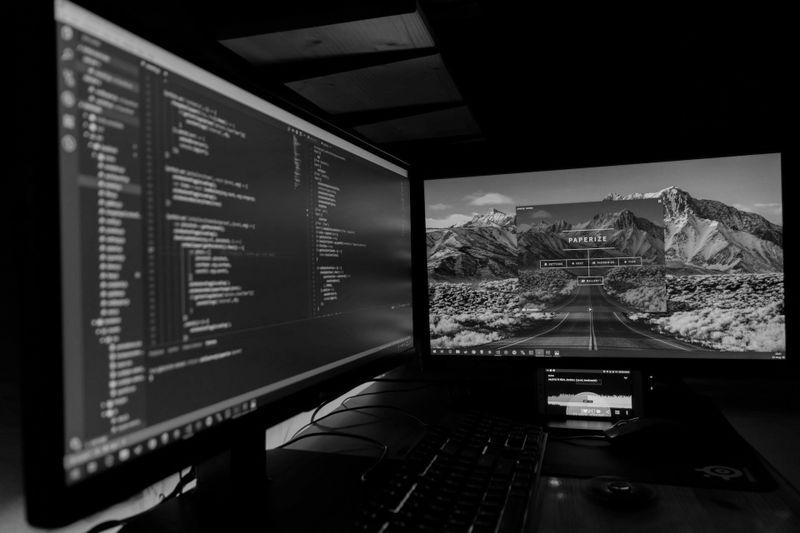 Zwei Computermonitore in Schwarz-Weiß, einer zeigt Code und der andere eine Berglandschaft mit einer Straße, Tastatur und Maus sind sichtbar.