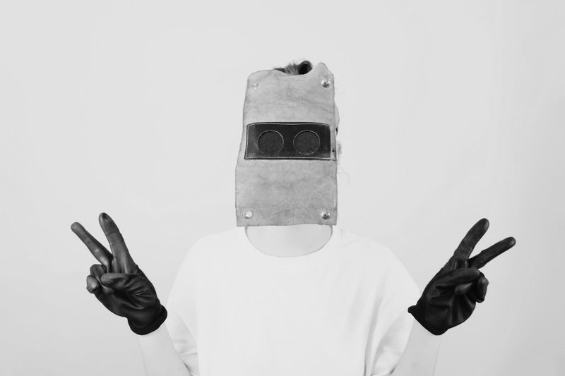Une personne portant un masque de soudage et des gants noirs, faisant des signes de paix avec les deux mains, sur une photo en noir et blanc.