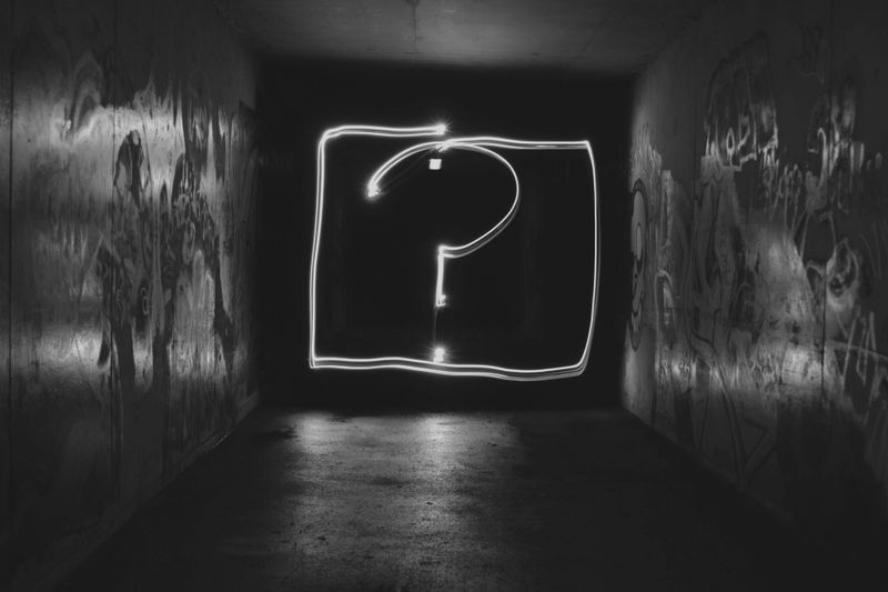 Un couloir sombre avec des murs couverts de graffitis présente un point d'interrogation lumineux dessiné avec de la lumière au centre.